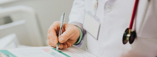 Doctor wearing a white coat holding a pen, writing on a clipboard.