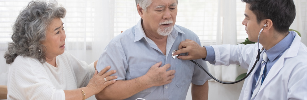 Doctor holding a stethoscope against an elderly patient's chest. The elderly patient holds one hand against his chest while an elderly female uses both hands to support the patient.